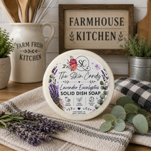 Load image into Gallery viewer, A round soap of the The Skin Candy Lavender Eucalyptus Solid Dish Soap sits on a checkered cloth with eucalyptus leaves and lavender sprigs. The background features a rustic wooden sign reading 'FARMHOUSE KITCHEN' and a ceramic pitcher labeled 'FARM FRESH KITCHEN'.