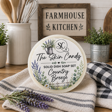 Load image into Gallery viewer, A round solid dish soap bar labeled 'The Skin Candy' with 'Country Breeze' scent, styled on a kitchen counter with lavender sprigs, a checkered cloth, and a rustic wooden sign in the background.

