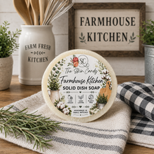Load image into Gallery viewer, A round package of soap from The Skin Candy. Farmhouse Kitchen Solid Dish Soap sits on a rustic wooden table with a striped towel, rosemary sprigs, a black-and-white check cloth, a ceramic utensil holder, and a framed sign reading 'Farmhouse Kitchen' in the background.
