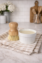 Load image into Gallery viewer, Wooden dish brush and white bowl on a kitchen counter with a towel and flowers in the background.
